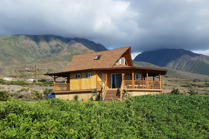 Wooden house with porch in green field, mountains in background under cloudy sky.